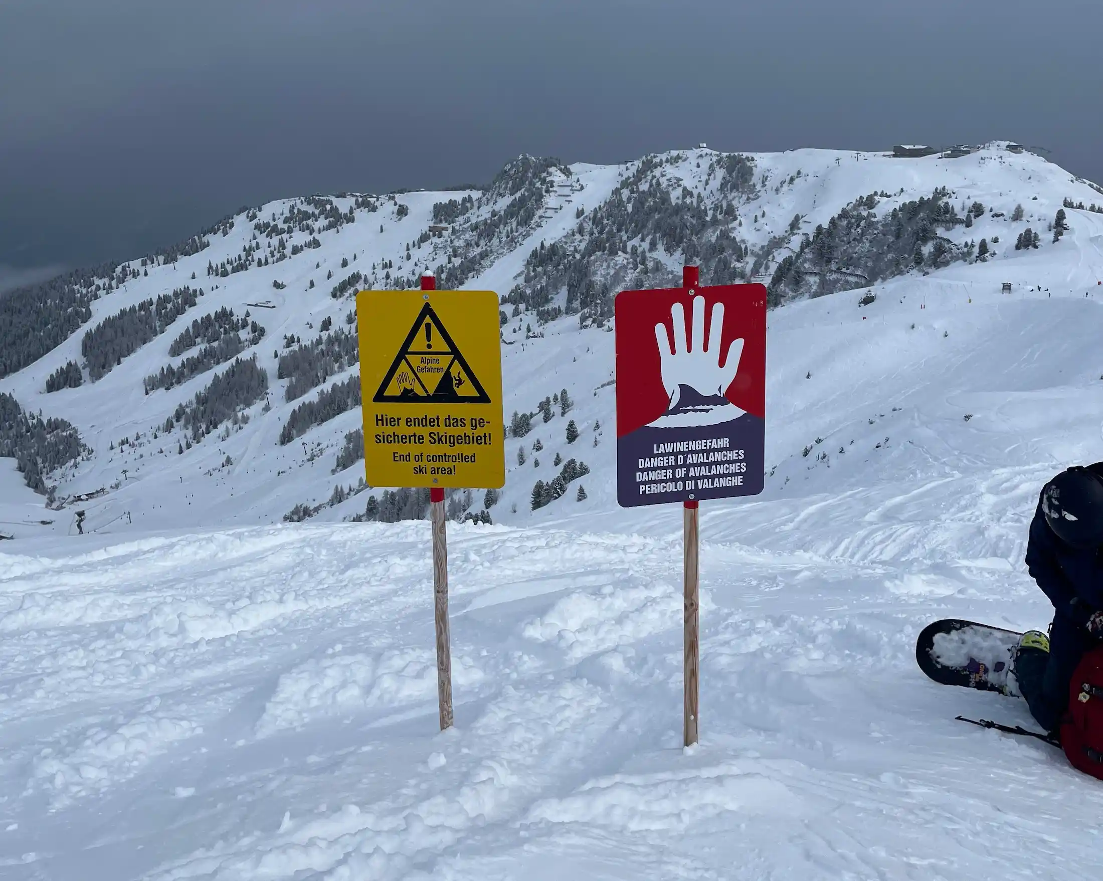 Avalanche warning signs at the edge of a controlled ski area in the snowy Alps, with mountains and fresh tracks in the background.
