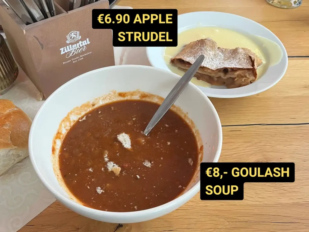 Bowl of goulash soup with bread on a wooden table, plus a plate of apple strudel with vanilla sauce, with price labels (€8 goulash soup and €6.90 apple strudel).