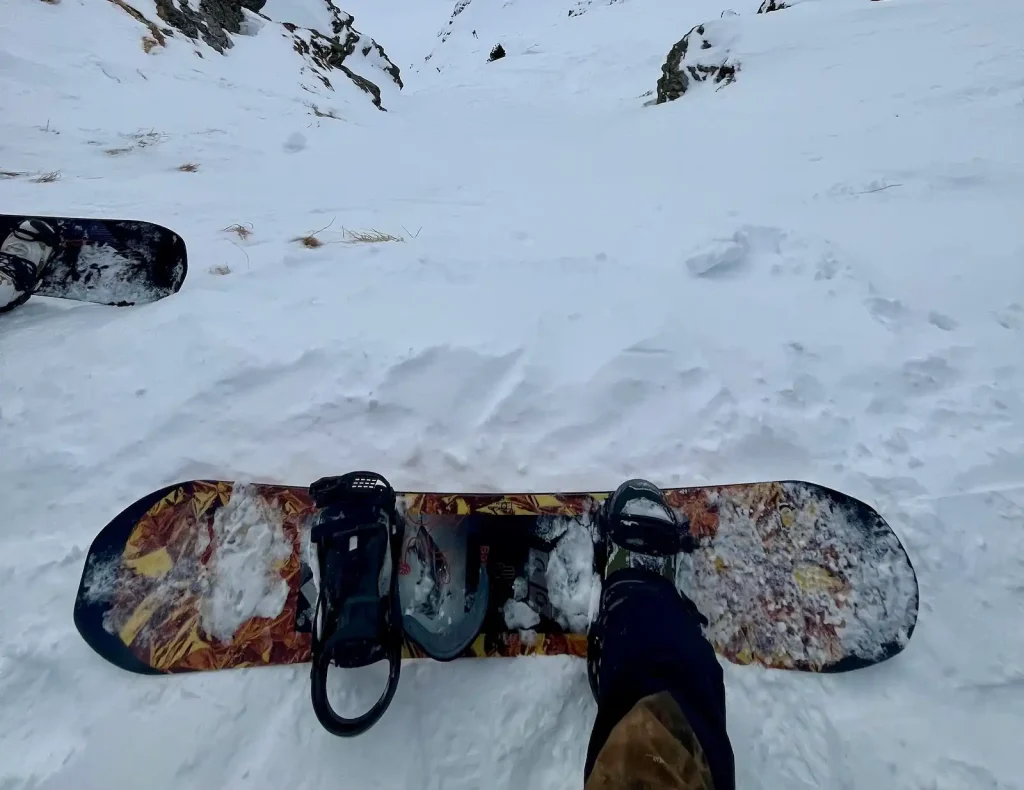 POV of a rider strapped into a Bataleon FunKink snowboard at the top of a snowy freeride drop, with a steep chute and rocky sides ahead.