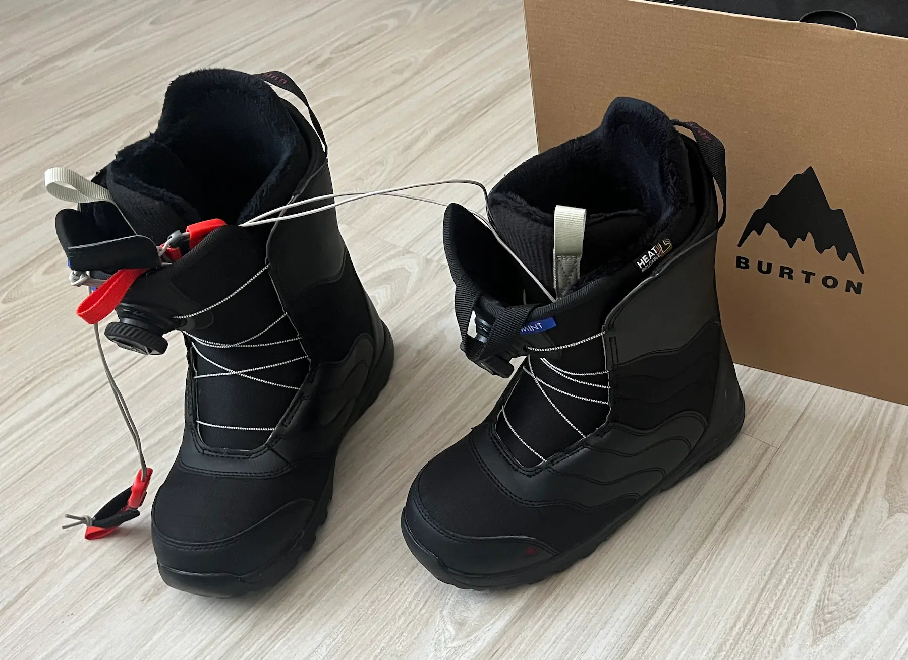 Two black Burton Mint BOA snowboard boots on a light wooden floor, one with the BOA cable loosened, next to a Burton-branded cardboard box.