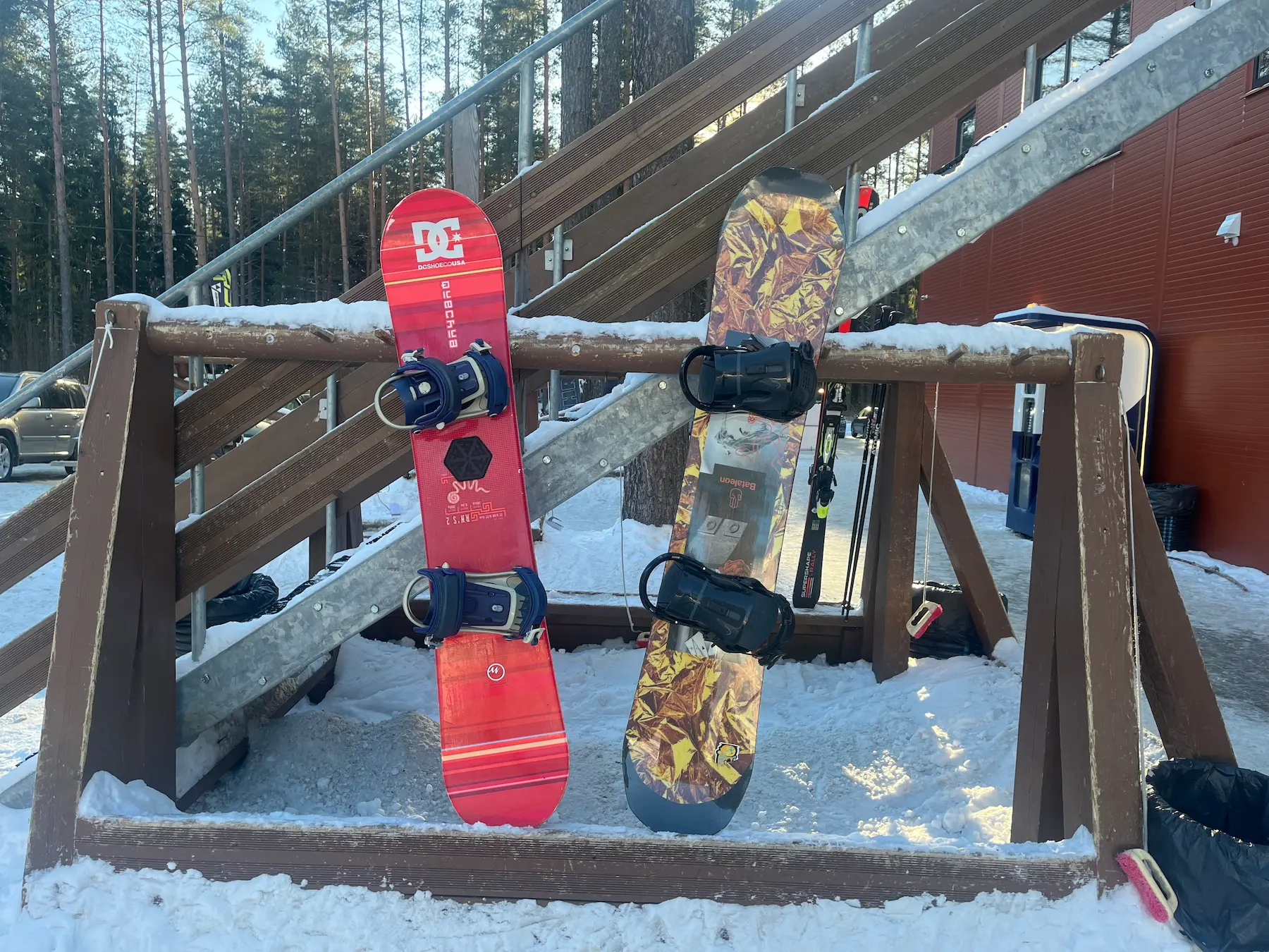 Two snowboards leaning against a wooden rack in the snow near a staircase: a red DC/Decathlon Wedze board and a Bataleon FunKink with black bindings.